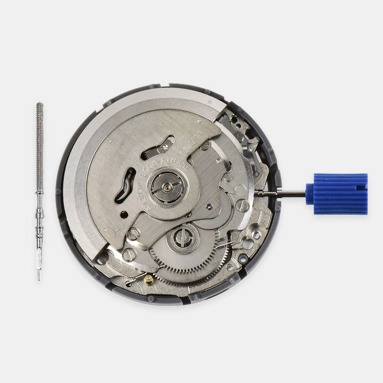 A watch movement case opened to show the internal mechanical gears and bridges, photographed on a light gray background. A small screwdriver is placed to the left, and a blue-handled tool is attached to the right.