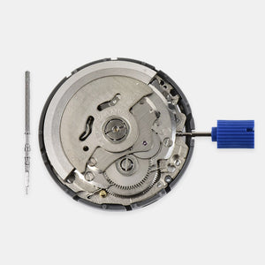 A watch movement case opened to show the internal mechanical gears and bridges, photographed on a light gray background. A small screwdriver is placed to the left, and a blue-handled tool is attached to the right.
