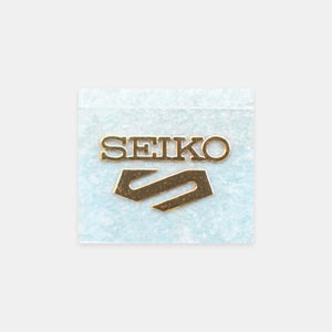 A gold-tone Seiko logo pin with the brand name and logo, displayed on a textured light blue square card against a light gray background.