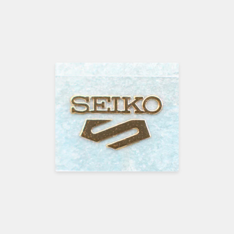 A gold-tone Seiko logo pin with the brand name and logo, displayed on a textured light blue square card against a light gray background.