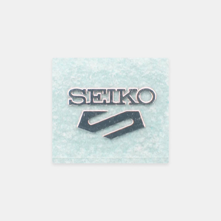 A square Seiko brand patch or emblem with the brand name in silver lettering and the Seiko logo below, set against a light icy blue textured background, centered on a light gray backdrop.