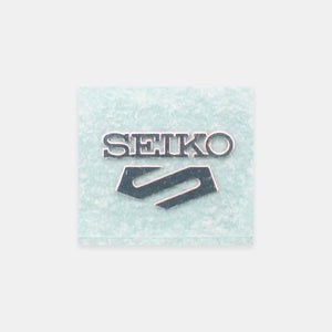 A square Seiko brand patch or emblem with the brand name in silver lettering and the Seiko logo below, set against a light icy blue textured background, centered on a light gray backdrop.