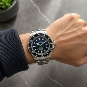 A wrist wearing a Seiko stainless steel dive watch with a black dial and black rotating bezel, photographed against a gray stone surface with a small plant in the background.