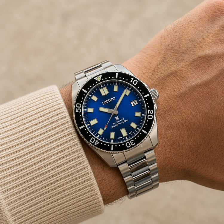 A Seiko stainless steel wristwatch with a blue dial and black bezel, shown worn on a person's wrist with a beige knit sleeve visible.