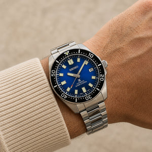 A Seiko stainless steel wristwatch with a blue dial and black bezel, shown worn on a person's wrist with a beige knit sleeve visible.
